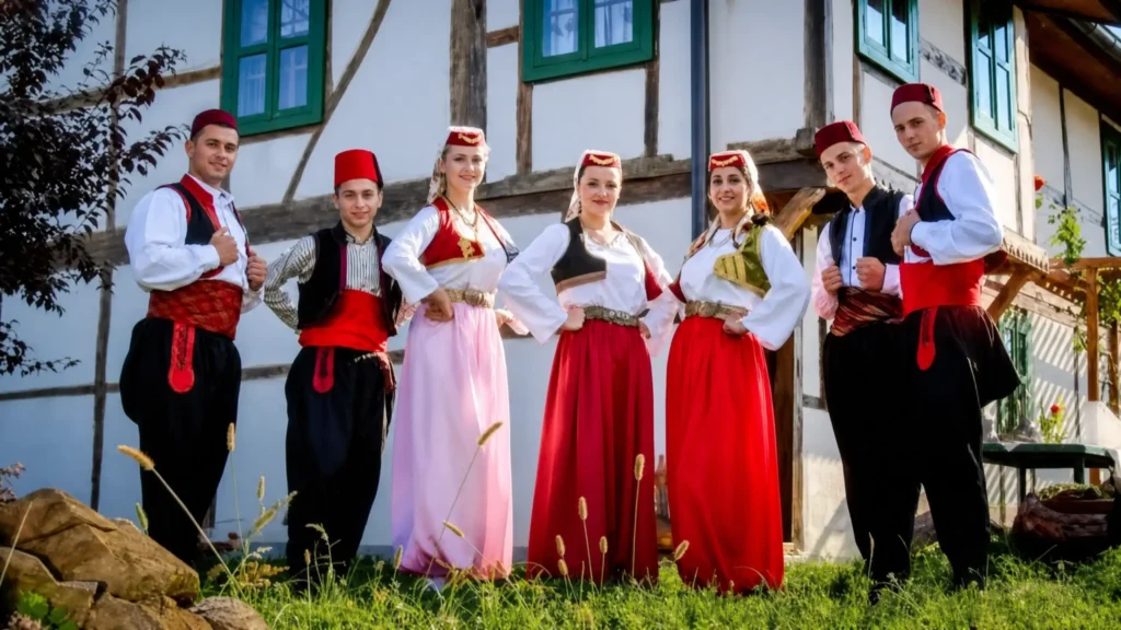 Realistična fotografija grupe mladića i djevojaka u autentičnim narodnim no&scaron;njama Bosne i Hercegovine, koji stoje ispred tradicionalne bijele kuće sa drvenim gredama na sunčan dan. Mu&scaron;karci nose bijele ko&scaron;ulje, crne prsluke i crvene fesove, dok djevojke nose vežene prsluke i duge, &scaron;arene suknje.